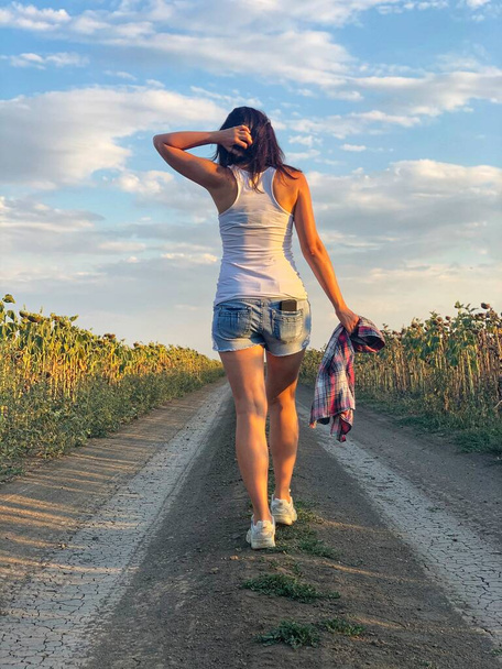 Chica joven en pantalones cortos de mezclilla y camiseta blanca está caminando a lo largo de la carretera rural. Vista desde atrás. - Foto, Imagen