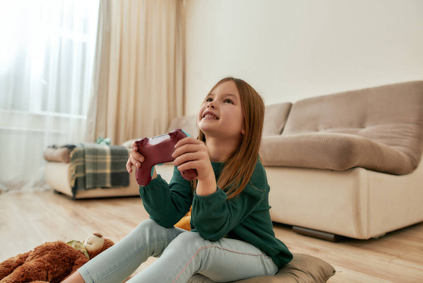 A small cute girl enjoy playing videogames smiling holding a gamepad while sitting on a floor - Foto, afbeelding