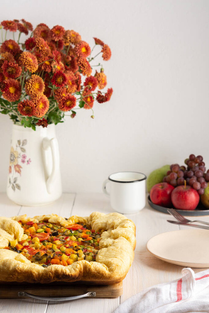 Pastete mit Gemüse, Hefeteig mit vorgekochtem Gemüse und im Ofen gebackenem Käse, weißer Tisch mit Pastete und Teller mit Gabel, Blumen in einer Vase - Foto, Bild