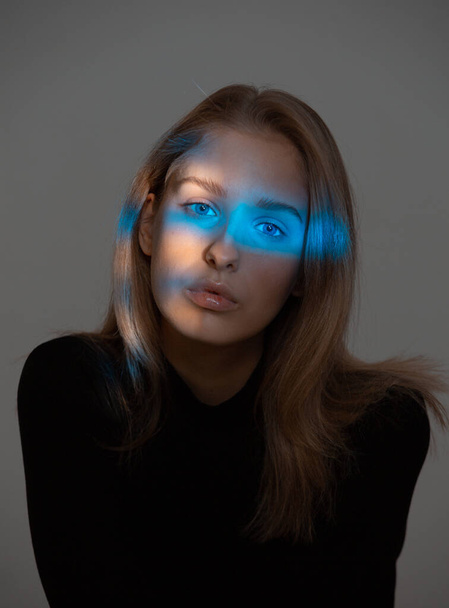 Portrait of young pretty girl with blue neon light on her face posing in studio on gray background - Photo, Image