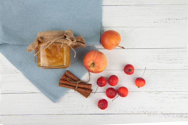 glass pot of jam, apples and cinnamon on blue napkin on white wooden background flat lay. Image contains copy space - Фото, зображення