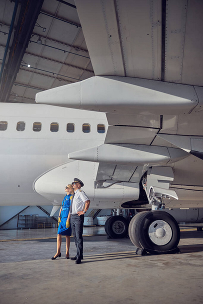 Hôtesse de l'air féminine avec capitaine regardant vers le ciel tout en se tenant au hangar de l'aéroport - Photo, image