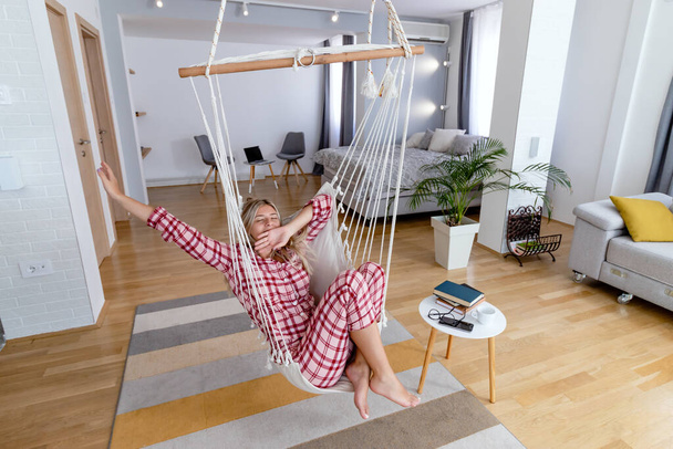 Beautiful young woman in pajamas yawning and stretching in a hanging hammock chair in a winter morning. Modern lifestyle and leisure concept. - Foto, afbeelding