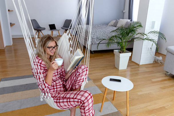 Hermosa joven en pijama leyendo un libro y disfrutando en una mañana de invierno con una taza de café en una silla de hamaca colgante. Estilo de vida moderno y concepto de ocio. - Foto, Imagen