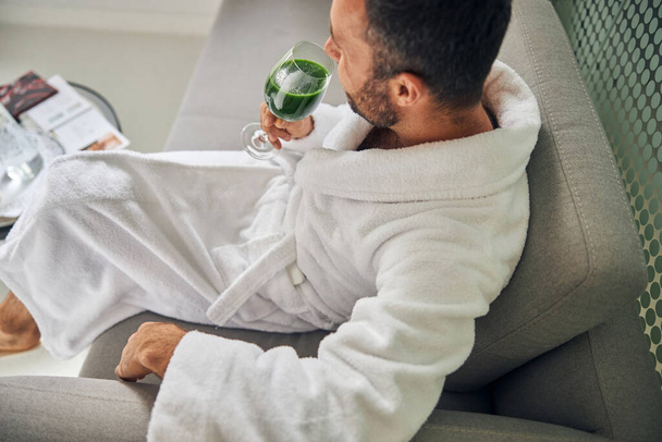 Бородатий молодий чоловік сидить на дивані і п'є зелену смужку
 - Фото, зображення