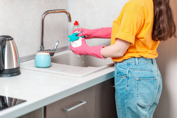 Trabajo doméstico. Una ama de casa con ropa casual y guantes de goma rosa está lavando una taza. Vista lateral. - Foto, Imagen