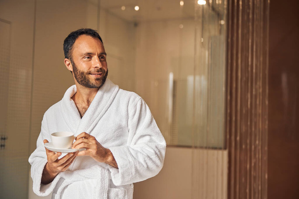 Joyful young man with coffee standing in spa salon - Foto, Bild