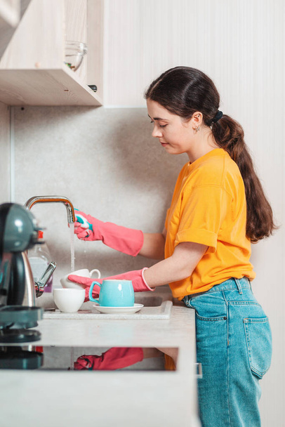 Hausarbeit. Eine hübsche Frau in lässiger Kleidung und rosa Gummihandschuhen spült Geschirr. Seitenansicht. - Foto, Bild