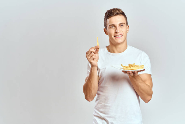 молодой парень с картошкой фри в коробке и в белой футболке на светлом фоне обрезанный вид - Фото, изображение