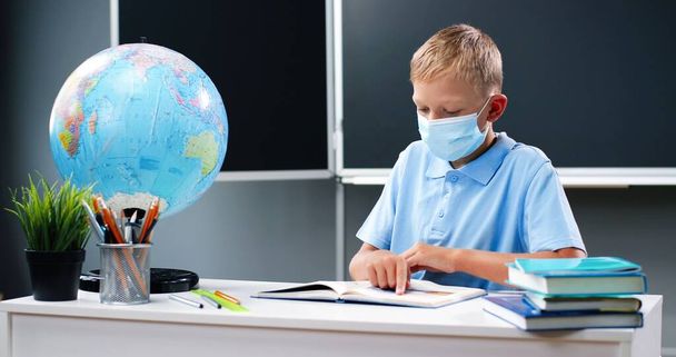 Small Caucasian teen boy in medical mask sitting at desk with world globe in school. Teenage schoolboy reading textbook and following with finger. Pupil learning in classroom. Coronavirus concept. - Foto, afbeelding