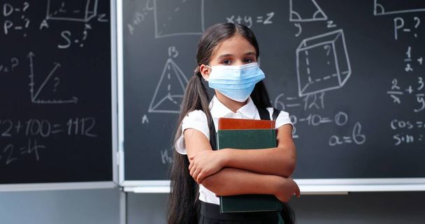 Portrait of cute small Caucasian pretty girl in medical mask and with backpack standing in classroom, looking at camera and holding textbooks. Little schoolgirl at blackboard in school. Pandemic time. - Zdjęcie, obraz