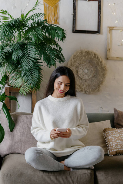 Dreamy and happy young adult girl sitting on couch, using modern smartphone, smiling wide and resting in living room at cozy home with plants - Fotografie, Obrázek