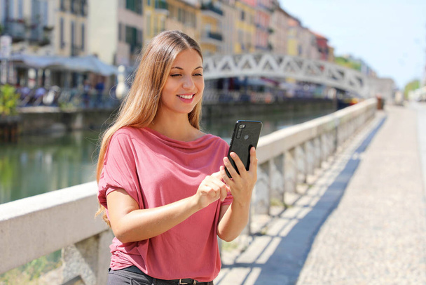 Χαμογελώντας νεαρή γυναίκα κυλιόμενα μηνύματα σε εφαρμογή smartphone για να συνομιλήσετε στο Μιλάνο, Ιταλία - Φωτογραφία, εικόνα