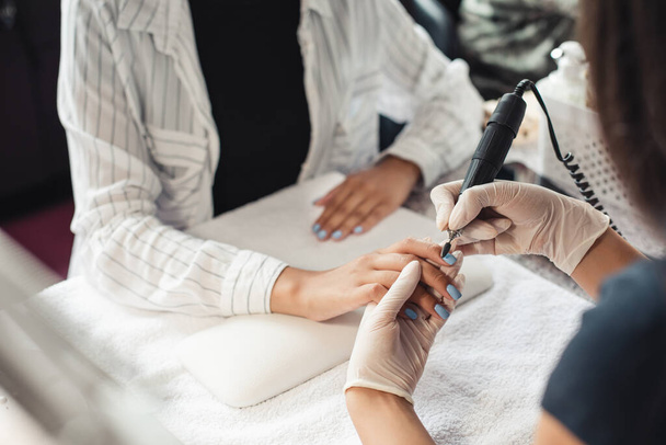 Beauty day and nail treatments. Professional manicure master in rubber gloves makes procedure removes varnish with an electric manicure device to african american client - Foto, Imagen