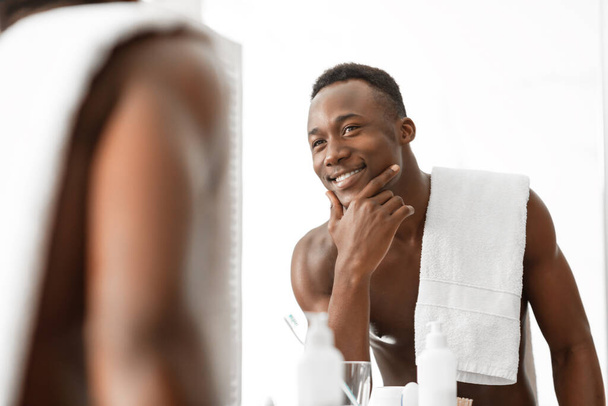 Knappe zwarte man op zoek naar zijn reflectie in de badkamer spiegel - Foto, afbeelding
