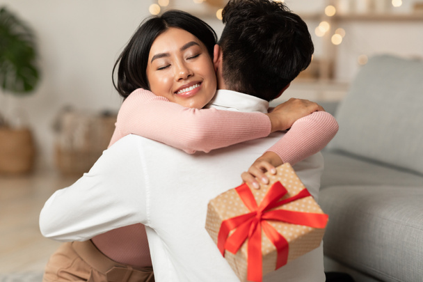 Joyeuse petite amie asiatique étreignant petit ami tenant boîte cadeau enveloppé à l'intérieur - Photo, image