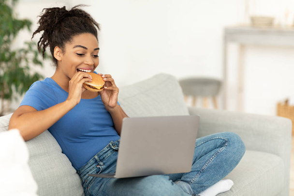 黒女は自宅でノートパソコンに座って不健康なバーガーを楽しむ - 写真・画像