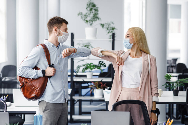 Modern greeting with colleague during coronavirus outbreak. Millennial guy and lady in protective masks came to work in morning and touch with elbows to say hello - Фото, изображение