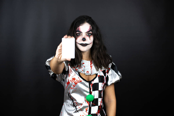 женщина в костюме клоуна Хэллоуина на изолированном черном фоне улыбается и держит белую карточку - Фото, изображение