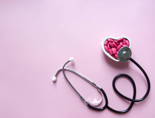 on a pink background vitamins of pink color in a plate a heart and a phonendoscope listens to heartbeat copy space  - Фото, изображение