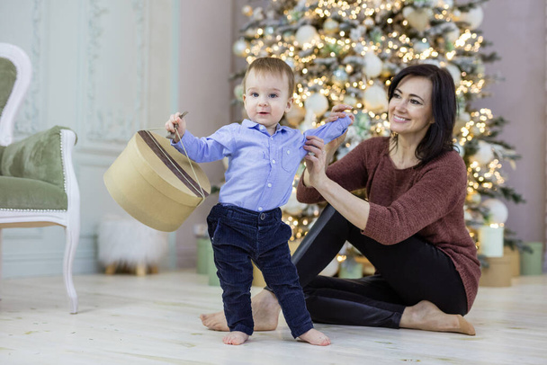 Atractiva mujer madura jugando con el nieto del bebé cerca del árbol de Navidad. Niño está llevando caja de regalo. - Foto, Imagen
