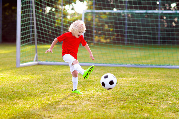 Dzieciaki grają w piłkę na boisku. Dzieci zdobywają gola podczas meczu. Mały chłopiec kopie piłkę. Biegające dziecko w koszulce drużyny i korkach. Szkolny klub piłkarski. Szkolenia sportowe dla młodych graczy. - Zdjęcie, obraz