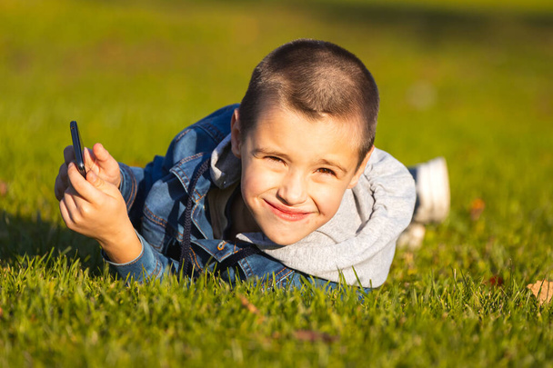 Un garçon joyeux dans une veste en jean se trouve sur une pelouse verte et joue avec un smartphone dans un parc de la ville. Concept du problème de la passion des enfants pour les jeux mobiles et les dessins animés - Photo, image