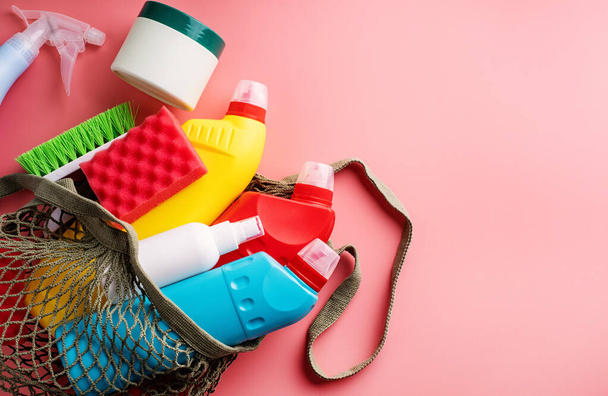 Προϊόντα καθαρισμού και καθαριότητας. Μπουκαλάκια υγιεινής και εργαλεία καθαρισμού σε οικολογική τσάντα πλέγματος σε ροζ συμπαγές φόντο - Φωτογραφία, εικόνα