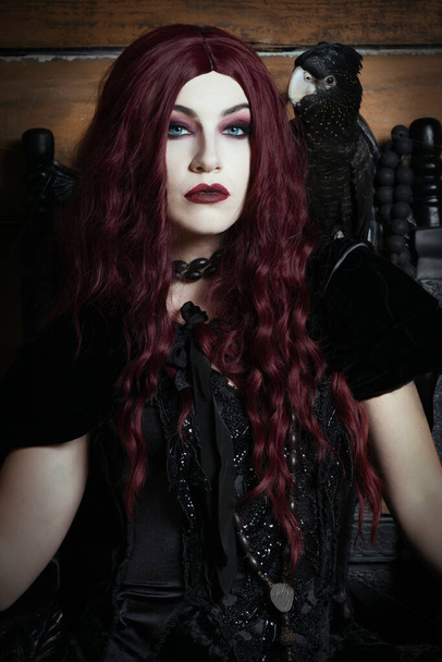Женщина, одетая как ведьма, сидящая на готическом троне с черным ара на плече - Фото, изображение