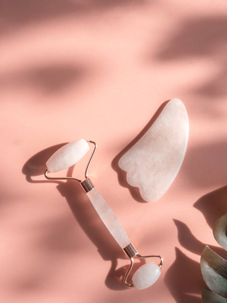 Massage pink natural quartz roller for the face skin care and facial massage with trendy sunny contrasting shadows. Gua Sha tools. Anti age, relaxation lifting at home and alternative medicine concept - Foto, Imagem