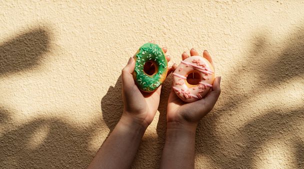 Das abgeschnittene Bild eines grünen und rosa Donuts wird von der Hand der Frau mit Licht und Schatten an die Wand gehalten. - Foto, Bild