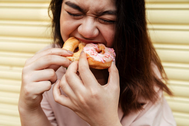 Woman eating two donuts in the same time while standing at the yellow shutter door. - Photo, Image