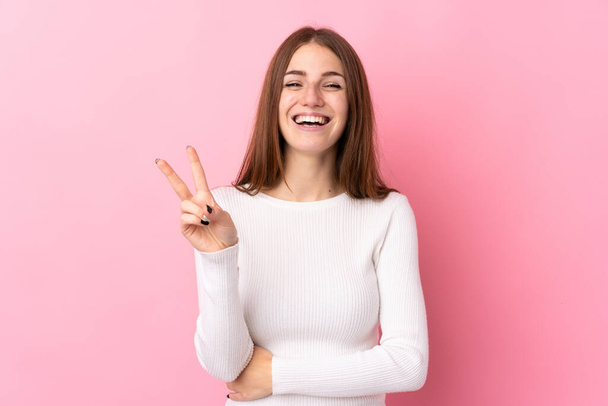 Junge Frau vor isoliertem rosa Hintergrund lächelt und zeigt Siegeszeichen - Foto, Bild