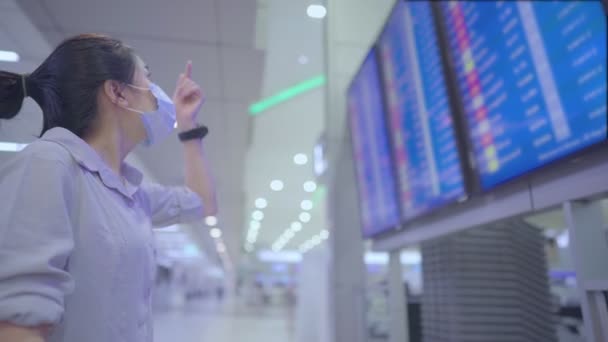 Young Asian female in white face mask checking time on wristwatch in front of airport flight time schedule board, at airport terminal, corona virus covid-19 pandemic, new normal social distancing - Footage, Video