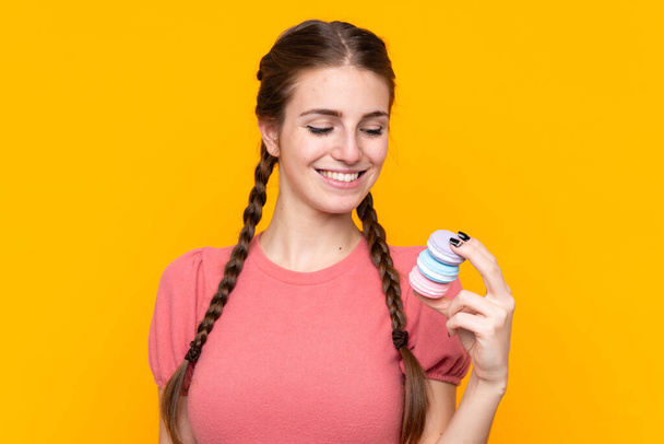 Young woman over isolated yellow background holding colorful French macarons and happy - Fotó, kép