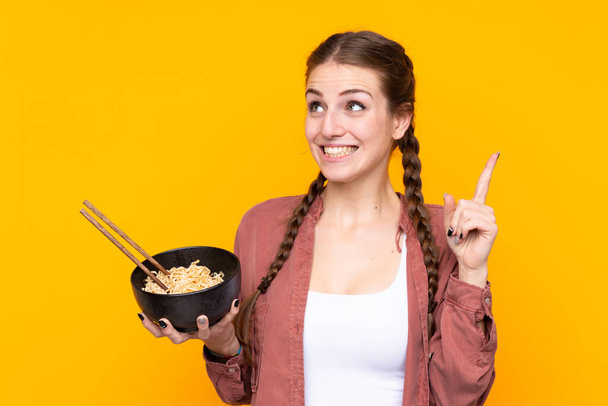 Young woman over isolated yellow background intending to realizes the solution while lifting a finger up while holding a bowl of noodles with chopsticks - Фото, изображение