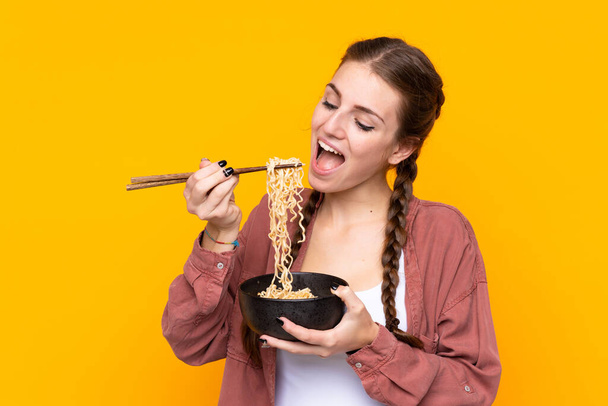 Jonge vrouw over geïsoleerde gele achtergrond met een kom noedels met eetstokjes en eten het - Foto, afbeelding