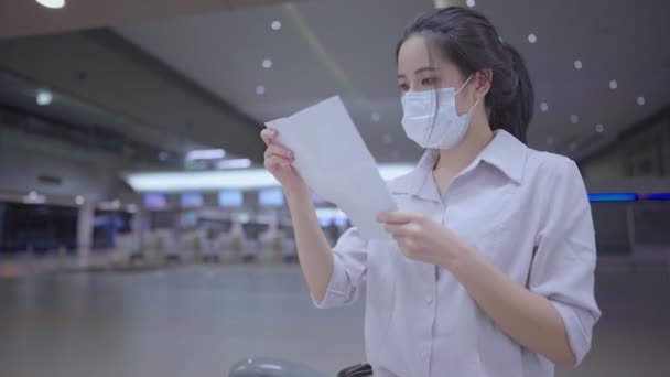 Azjatycka młoda kobieta nosi maskę medyczną czytaną na karcie podróży, sprawdza czas odprawy, wózek bagażowy, pusty terminal lotniska podczas pandemii dystansu społecznego - Materiał filmowy, wideo