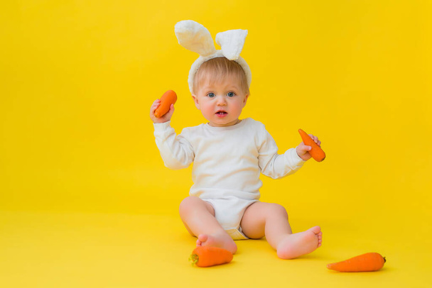 Bebé en un traje de cuerpo blanco con orejas de conejo en la cabeza come zanahorias, se sienta sobre un fondo amarillo con verduras. bebé en forma de conejito de Pascua, espacio para el texto - Foto, imagen