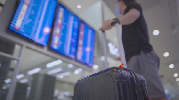 Азіатський чоловік носить маску перед екраном календаря польоту в аеропорту, реєстраційний лічильник на багажнику, на терміналі аеропорту, пасажир і валіза з низьким кутом огляду вгору. - Кадри, відео