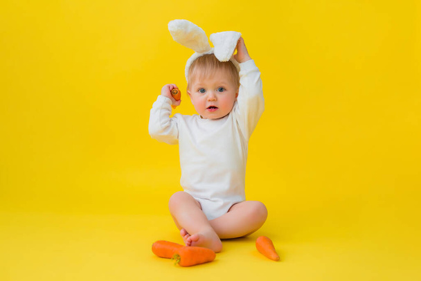 Dítě v bílém obleku s králičíma ušima na hlavě jí mrkev, sedí na žlutém pozadí se zeleninou. dítě v podobě velikonočního zajíčka, prostor pro text - Fotografie, Obrázek
