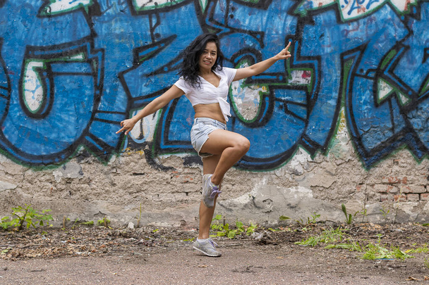 A cheerful young and dark-skinned girl in a white top and blue shorts stands against the background of a wall with graffiti with a raised leg and arms spread out to the sides - Photo, Image