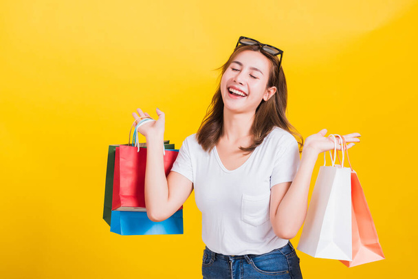 Asian Thai portrait happy beautiful cute young woman smiling stand with sunglasses excited holding shopping bags multi color close eyes, studio shot isolated yellow background with copy space - Fotografie, Obrázek