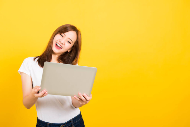 Asian happy portrait beautiful cute young woman teen smile standing wear t-shirt hold laptop computer and excited celebrating success looking to camera, studio shot yellow background with copy space - Foto, Bild