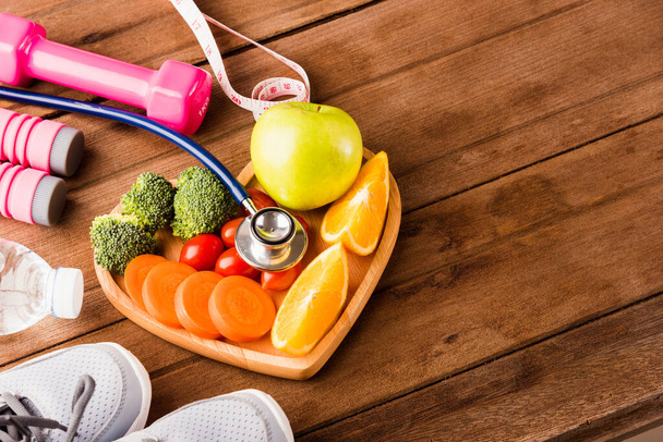 Top view of fresh fruits and vegetables in heart plate wood (apple, carrot, tomato, orange, broccoli) and sports equipment and doctor stethoscope on wooden table, Healthy lifestyle diet food concept - Photo, image