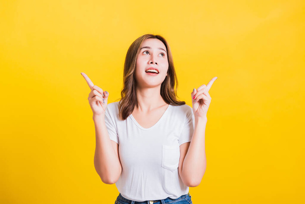 Asian Thai happy portrait beautiful cute young woman standing wear white t-shirt makes gesture two fingers point upwards above looking above, studio shot isolated on yellow background with copy space - Фото, изображение