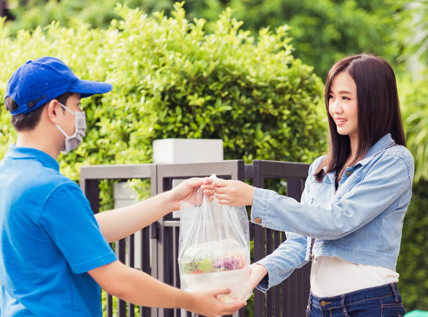 Asian young delivery man wear face mask he making grocery service giving rice food boxes plastic bags to woman customer receiving door at house after pandemic coronavirus, Back to new normal concept - 写真・画像