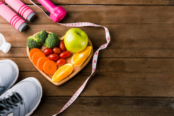 Top view of fresh fruits and vegetables in heart plate wood (apple, carrot, tomato, orange, broccoli) and dumbbells, sport shoes sports equipment on wooden table, Healthy lifestyle diet food concept - 写真・画像