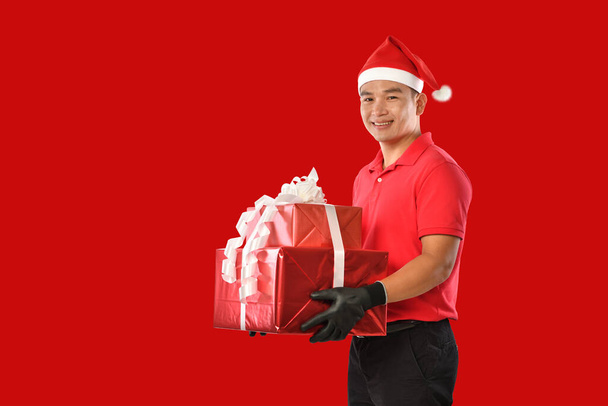 Ευτυχισμένος νεαρός Ασιάτης ντελιβεράς με κόκκινη στολή, χριστουγεννιάτικο καπέλο κουβαλάει κουτιά με δώρα στο κόκκινο φόντο κατά τη διάρκεια των γιορτών των Χριστουγέννων - Φωτογραφία, εικόνα