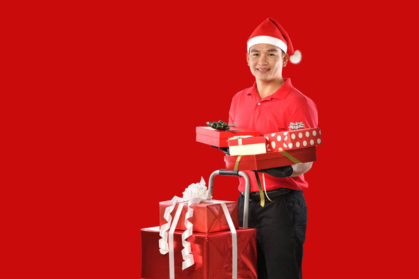 Glücklicher junger asiatischer Auslieferer in roter Uniform, Weihnachtsmütze trägt Schachteln mit Geschenken im praktischen roten Hintergrund während der Weihnachtsfeierlichkeiten - Foto, Bild